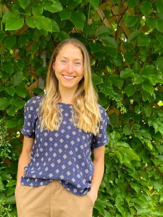 A smiling woman with long blonde hair standing in front of greenery