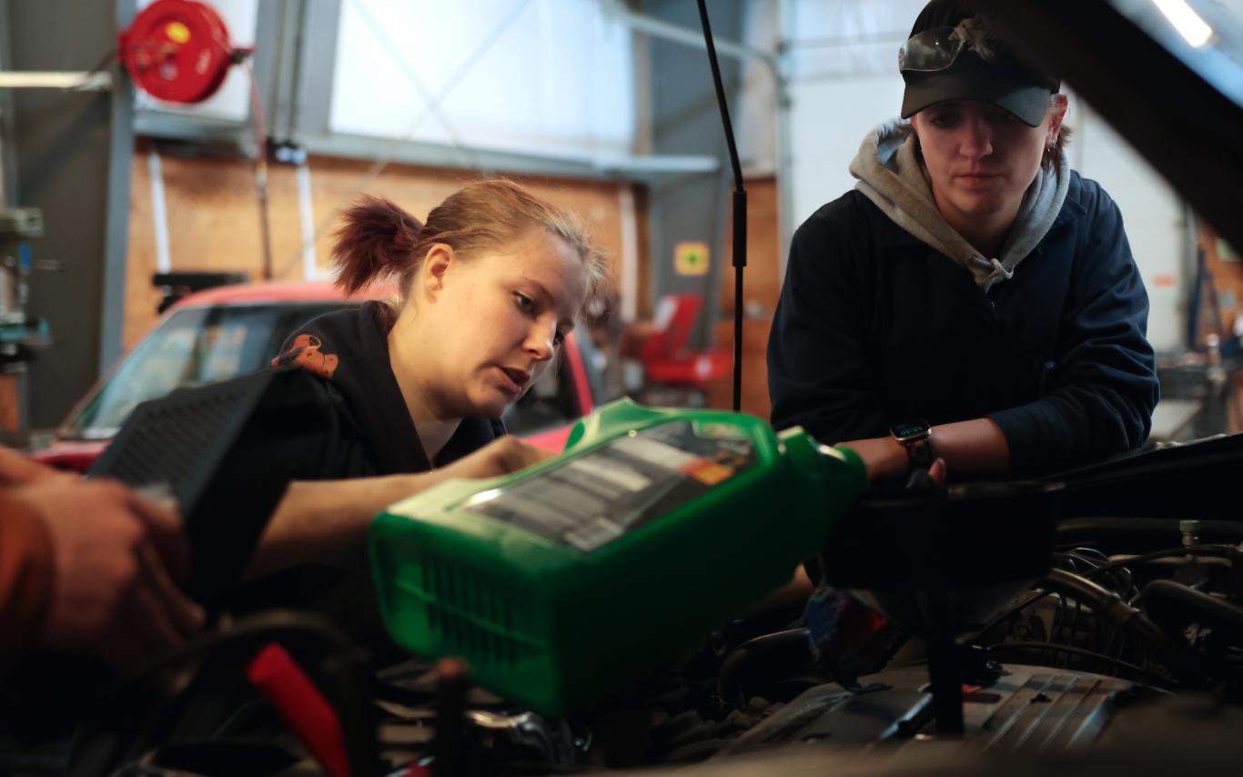 A young woman working on a car while a young man watches. 